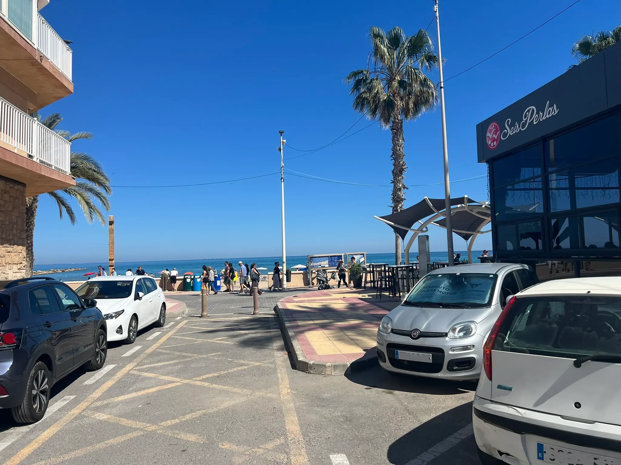 A street in El Campello leading toward the Mediterranean Sea. The scene shows parked cars, a palm tree, and the Seis Perlas restaurant on the right, with people walking along the beachfront promenade in the background.